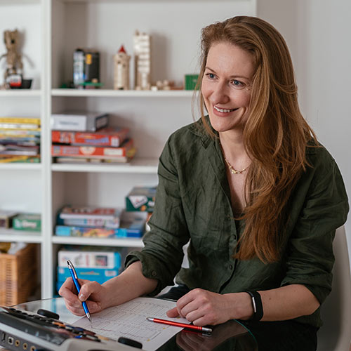 Portrait of woman with long hair and darj green t-shirt smiling sitting near the table with the pencil in her hand