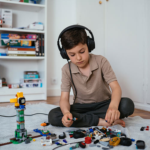 Boy with short dark hair is sitting on the ground with Tomatis headphones and playing Lego