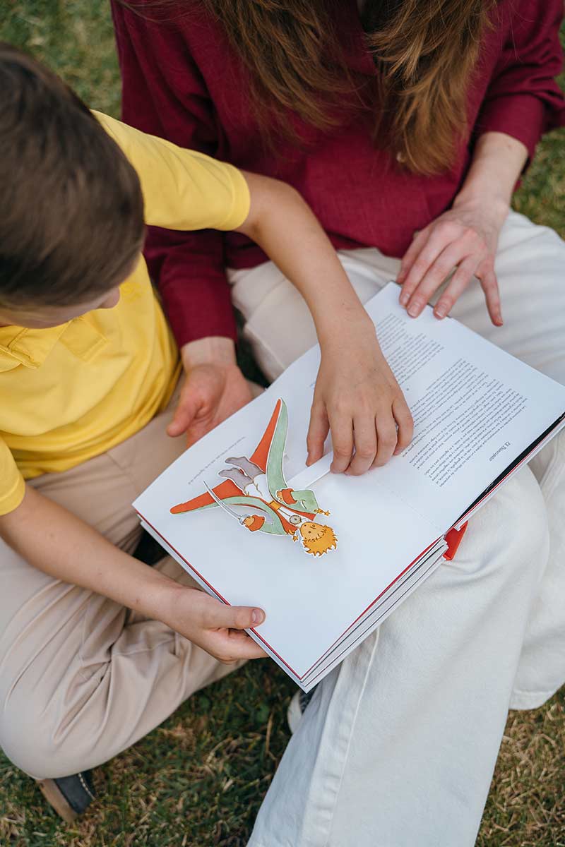 Mother and child sitting together outdoors, reading a colorful illustrated book, with the mother gently guiding the child’s attention to the page.