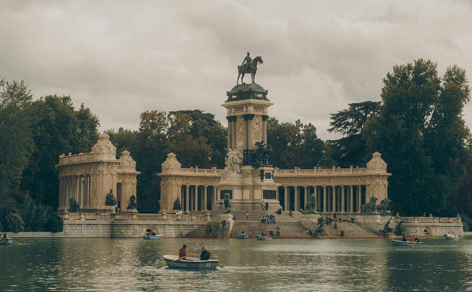 Wide view of a grand stone monument with an equestrian statue at its center, reflected in a calm lake where people are rowing small boats, surrounded by trees under a cloudy sky in Madrid