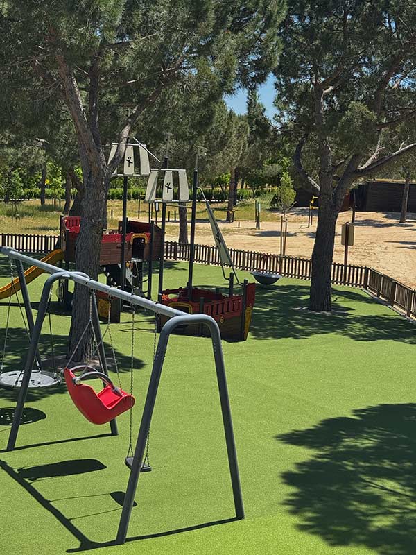 Children’s playground in Parque Juan Carlos I with swings and a pirate ship themed play structure, set on green safety flooring and surrounded by pine trees on a sunny day